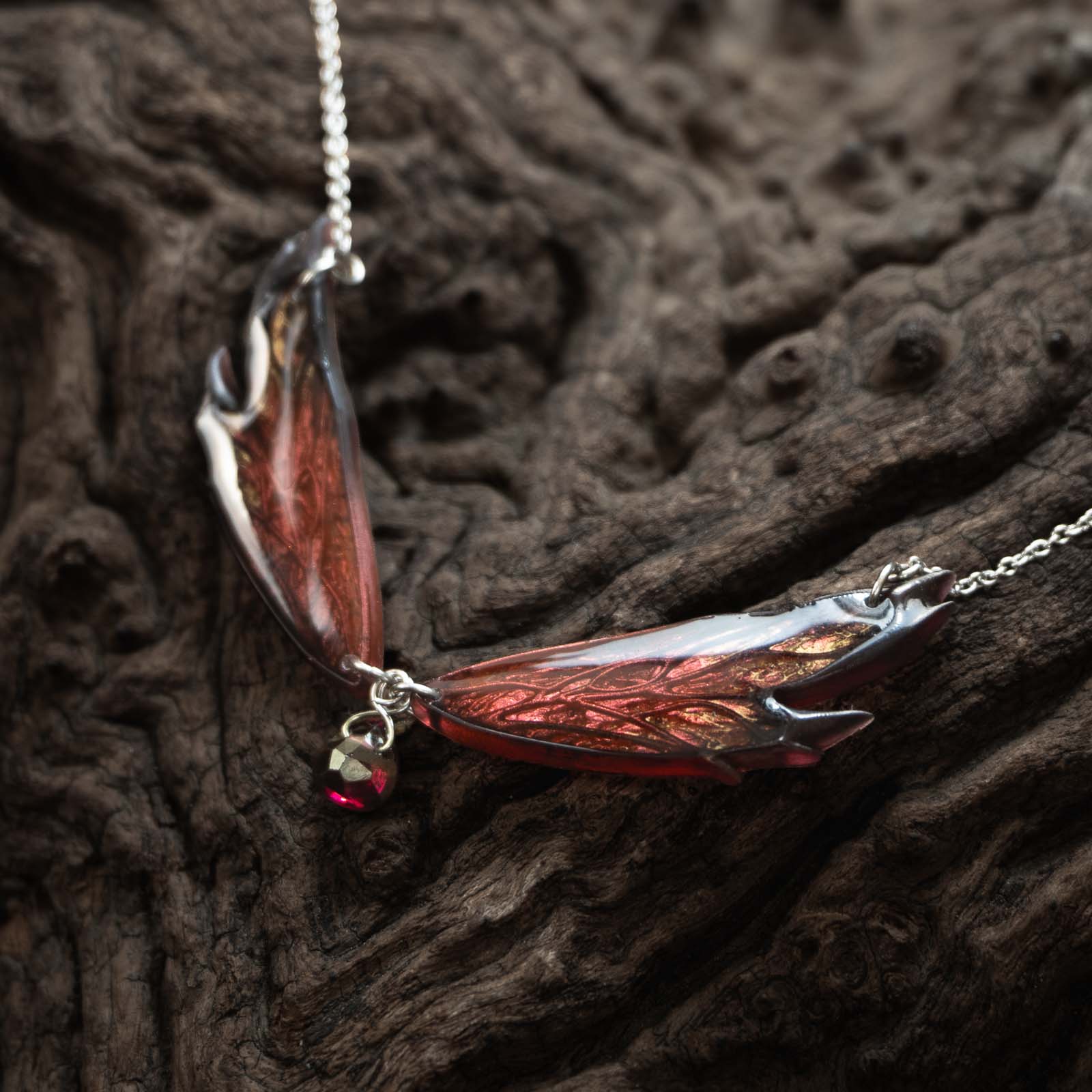 Collier Morgane Élégance - Sur bois Collier Morgane Élégance posé sur une souche de bois sombre, avec ses ailes de fée rouge cuivré aux contours noirs et son pendentif en grenat central.