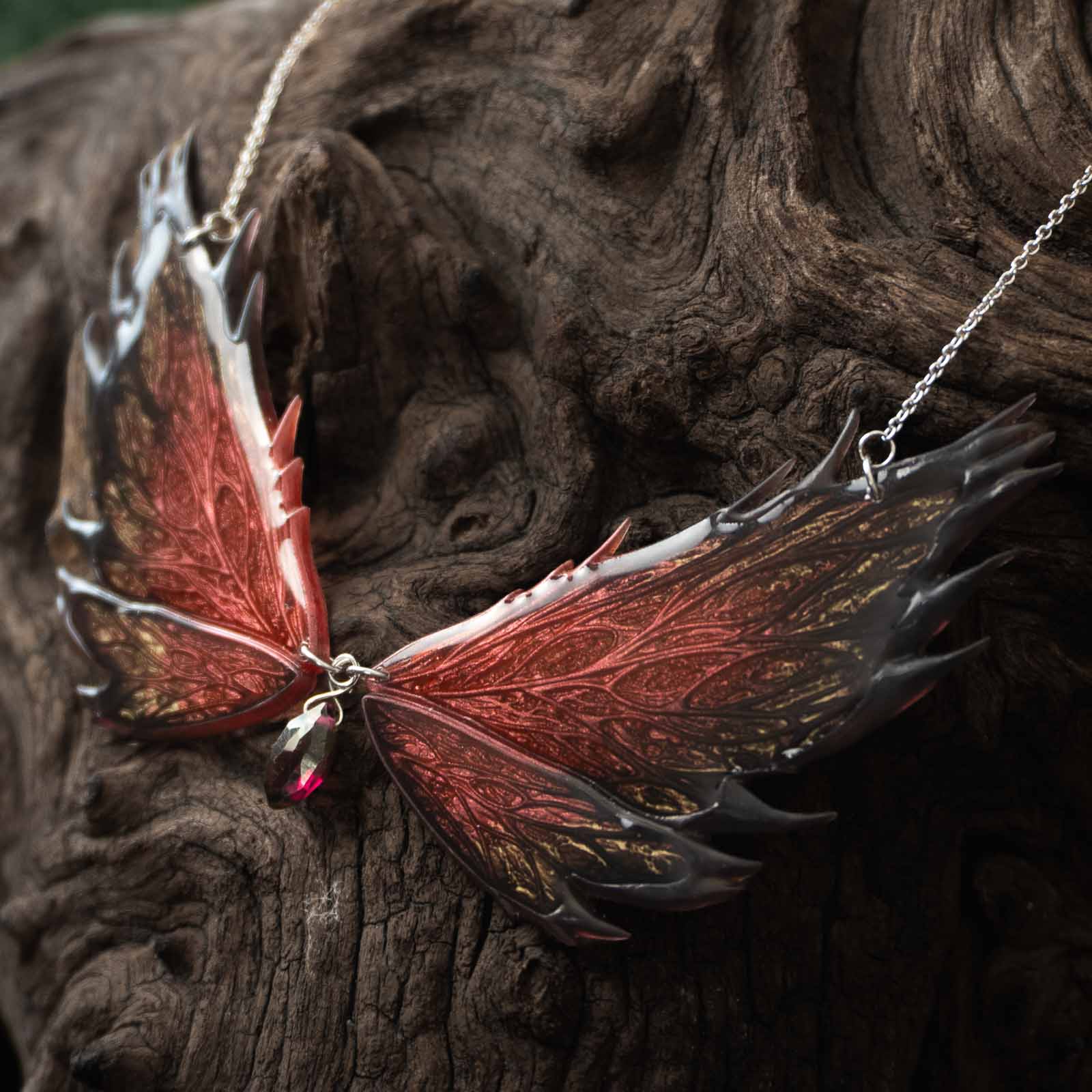 Collier Morgane Prestige posé sur du bois ancien, ses deux ailes de fée rouge sombre et noir aux reflets or déployées.